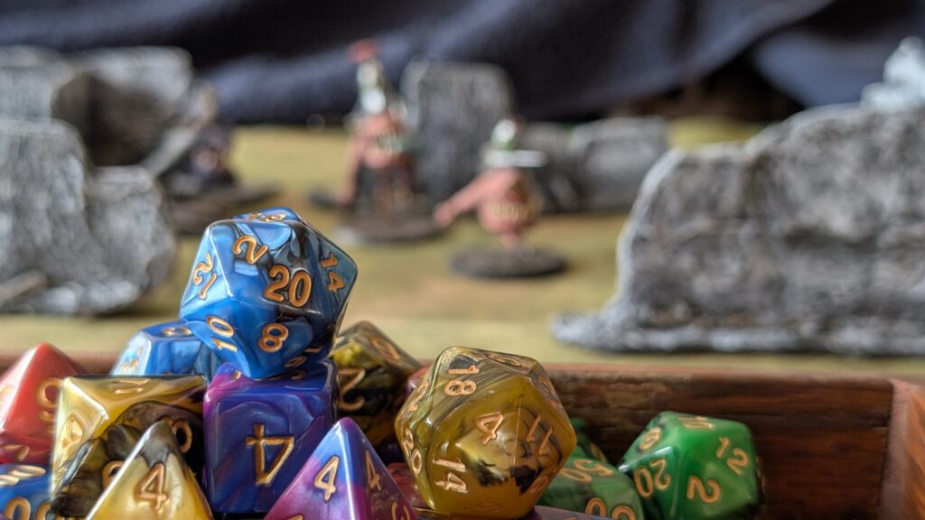 Dnd Dice on table top terrain Set of polyhedral Dungeons & Dragons dice resting in a leather dice tray, with blurred tabletop terrain and fantasy miniatures in the background.